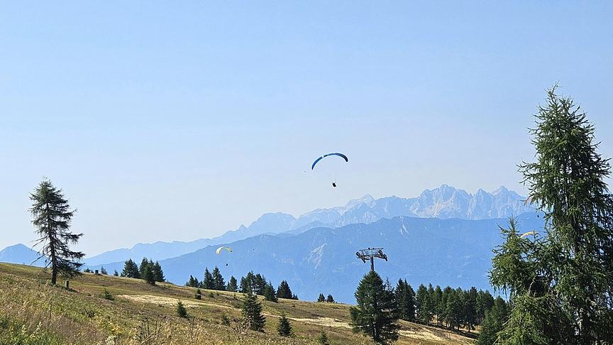 Eine weite Berglandschaft unter klarem blauem Himmel, in der mehrere Gleitschirmflieger über einer hügeligen, teils bewaldeten Wiese vor einer imposanten, im Hintergrund aufragenden Gebirgskette schweben.