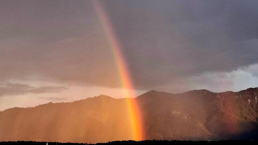 Ein Regenbogen zieht sich bei milchiger Regenstimmung vom Himmel zum Boden vor einem Bergpanorama über dem eine dichte Wolkendecke hängt.