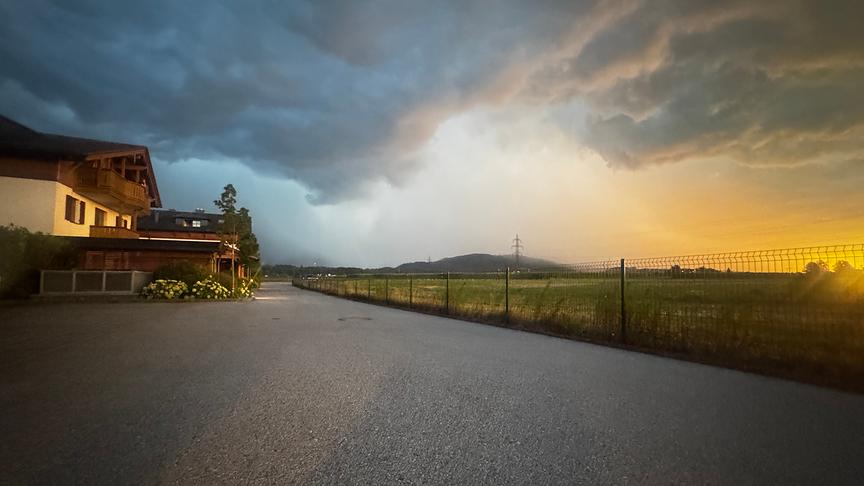 Eine weitwinklige Außenaufnahme zeigt eine asphaltierte Straße, die im Vordergrund beginnt und nach hinten links verläuft, unter einem dramatischen, zweigeteilten Gewitterhimmel.