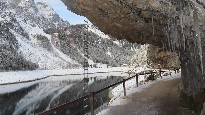© Rudolf Kößl Ein Weg führt neben einem von Bergen ungebenen See vorbei und unterhalb eines von Eiszapfen behangenen Felsvorsprung hindurch.