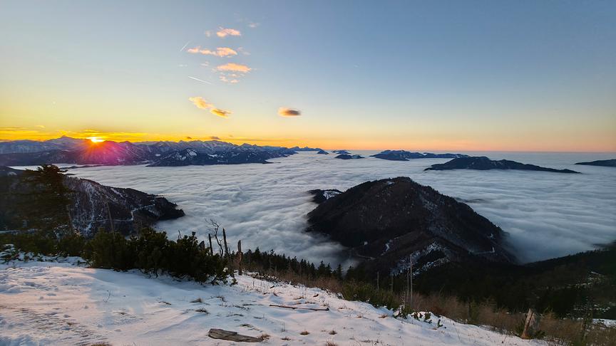© Christian Hahn Die Sonne geht über einer Wolkendecke und hinter einer Bergkette warmleuchtend unter.