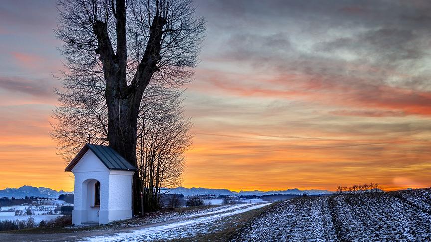 Leuchtend oranges Abendrot über den Bergen, im Vordergrund eine kleine Kapelle mit stattlichem Baum neben einem Feldweg