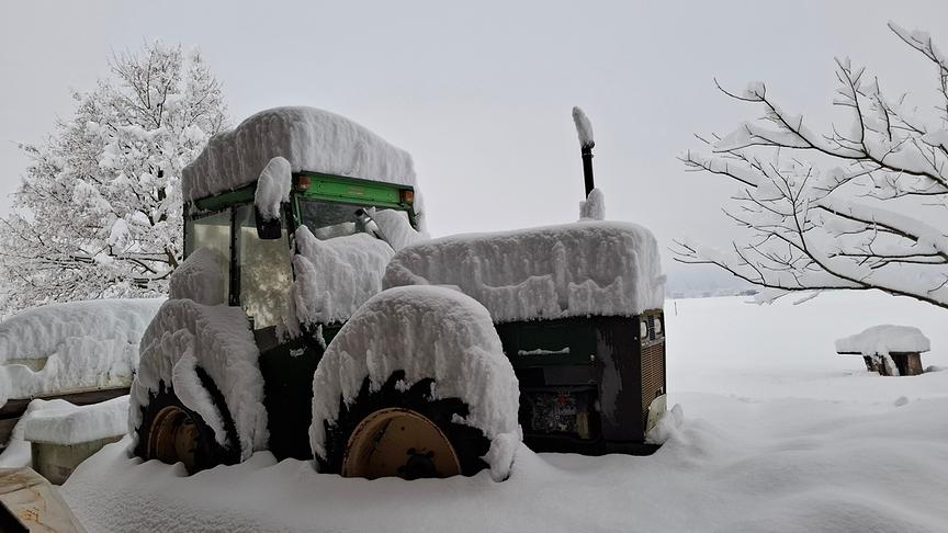 Schnee türmt sich centimeterhoch auf den waagerechten Flächen eines Traktors. Auch die Landschaft darumherum ist stark verschneit.