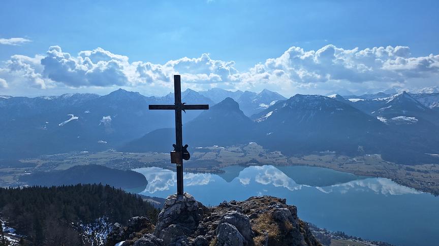Bergpanorama mit blauem Himmel und Wolken, fotagrafiert von einem Gipfel mit Kreuz, im Tal ein ruhiger See, indem sich Berg und Wolken spiegeln.