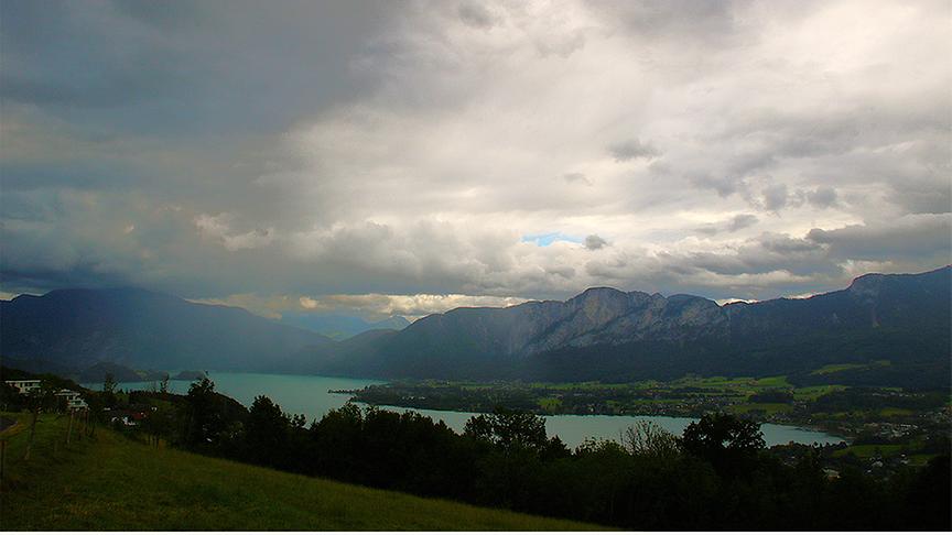 Wolkendecke über See und Bergen mit einem Schleier, der sich Richtung Boden zieht.