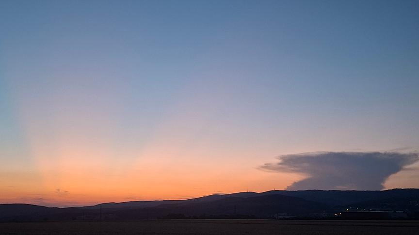 Silhouette einer flachen Hügellandschaft, über der sich die letzten warmen Sonnenstrahlen mit orangem Licht an den ansonsten schon dämmernden Himmel erstrecken. Eine Wolke ergänzt die Szenerie.