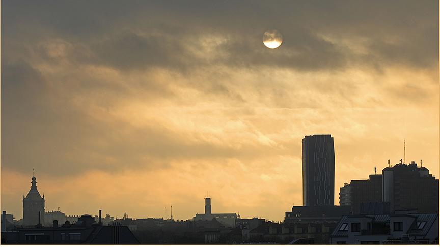 Die Sonne leuchtet orange durch schleiernde Wolken hindurch über dem Stadtpanorama. 