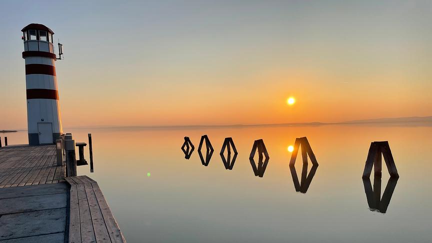 Sonnenuntergang über stillem See von einem Steg mit Leuchtturm aus gesehen