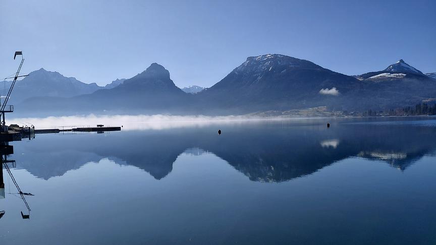 Stiller See über dem ein Streifen Nebel hinweg zieht, die Bergkulisse im Hintergrund und der blaue Himmel spiegeln sich im See