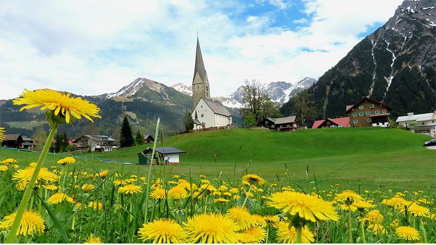 Im Vordergrund: Löwenzahn in einer Wiese, im Hintergrund: Kirche, Siedlung und hohe Berge bei bewölktem, aber hellem Himmel.