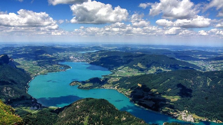 Im Tal schlängelt sich ein dunkel-türkiser See durch bewaldete Berge. Die Schönwetterwolken am Himmel werfen große Schatten auf die Landschaft.