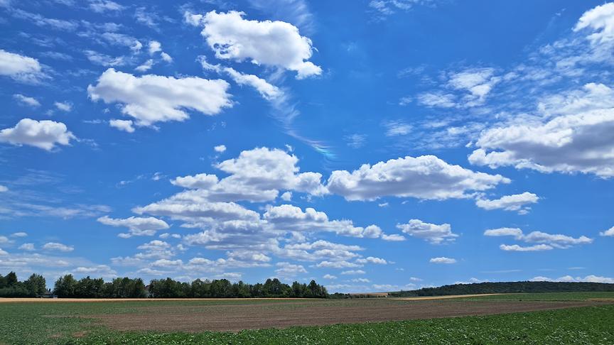 © Brigitte Lehner Vereinzelte Schönwetterwolken über eine weiten, flachen Landschaft mit Acker und Wäldern. Mittig im Bild ist eine Schlierenwolke zu sehen, die in den Regenbogenfarben erstrahlt.