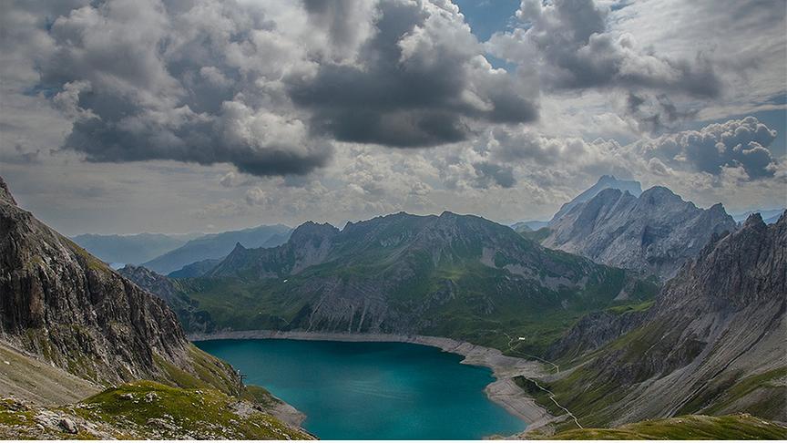 Felsige und teils noch grüne Bergkulisse mit einem türkisblauen See in der Mitte. Der Himmel ist gefüllt mit Wolken.