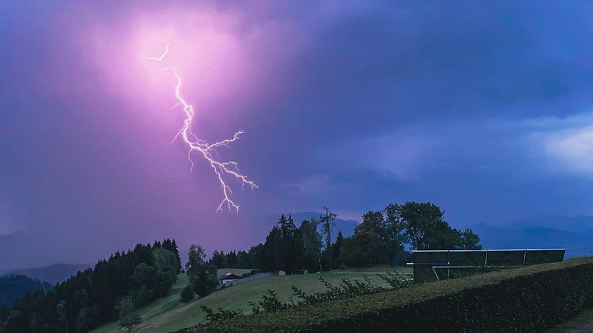 Heller, violettleuchtender Blitz über einer Hügellandschaft am dunklen Himmel.