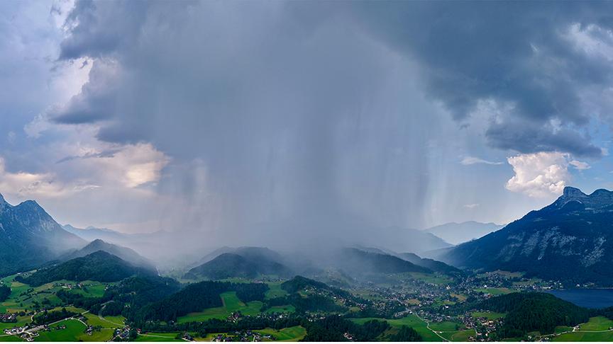 Bergige Landschaft mit Bäumen und Häusern sowie einem See. Im Fokus ist eine Gewitterzelle, die sich in deutlichen Schlieren zu Boden zieht. 
