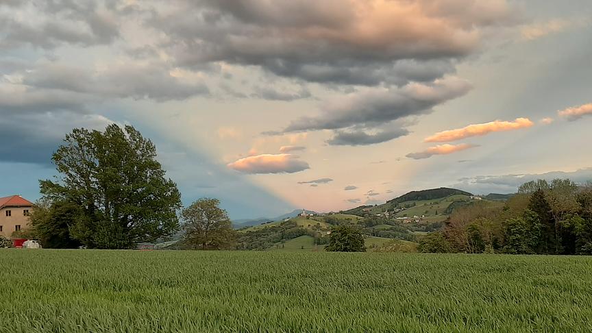 Hügelige Landschaft mit Siedlungen. Darüber Himmel mit Wolken, die Sonne stahlt einen schmalen Lichtkorridoren hinter einem Berg hervor.