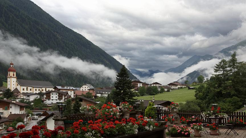 © Horst Fankhauser Ein malerisches Bergdorf in den Alpen, vermutlich Neustift im Stubaital, unter einem dramatisch bewölkten Himmel. Im Vordergrund blühen leuchtend rote Geranien auf einer Terrasse. Dahinter schmiegen sich traditionelle Häuser mit einer markanten Kirche mit rotem Zwiebelturm in ein grünes Tal, während dichte Nebelschwaden an den bewaldeten Berghängen hängen.
