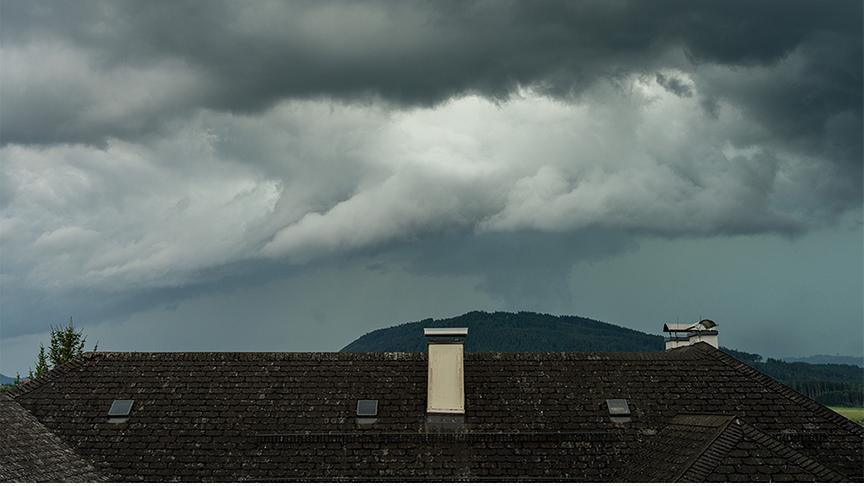 Dunkle, imposante Wolken türmen sich am grauen Himmel über einem Häusedach. Im Hintegrund ist ein Hügel, zu dem sich eine Wolke sichtbar hinunterzieht.