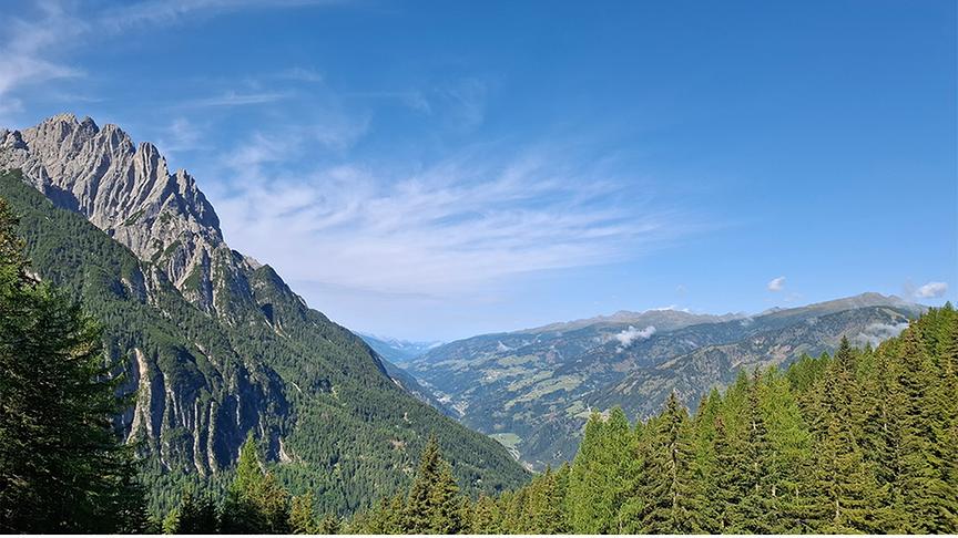 Bergige, waldbewachsene Landschaft unter blauem Himmel mit kunstvollen Schleierwolken.