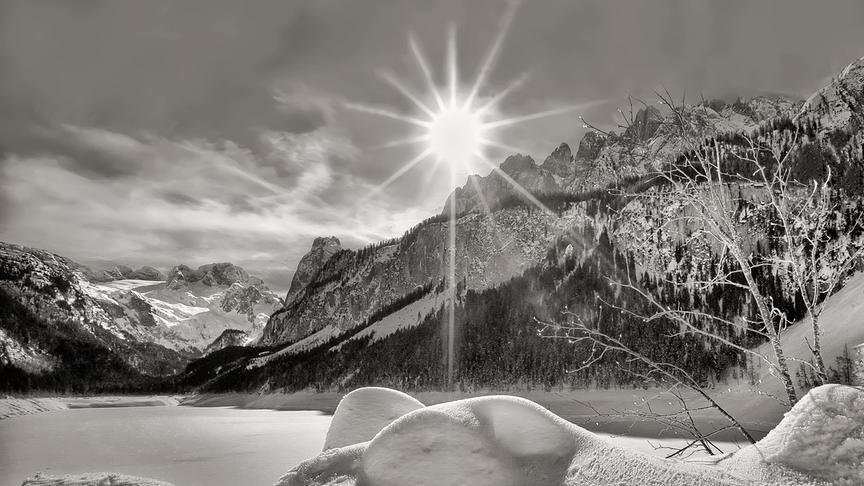 Schwarz-weiß Bild eines verschneiten Sees umgeben von felsigen Bergen und reiner strahlenden Sonne im Zentrum