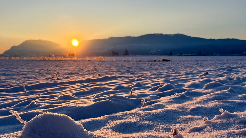 Nahaufnahme vom verschneiten, unebenem Boden mit Bergen und Sonnenaufgang im Hintergrund