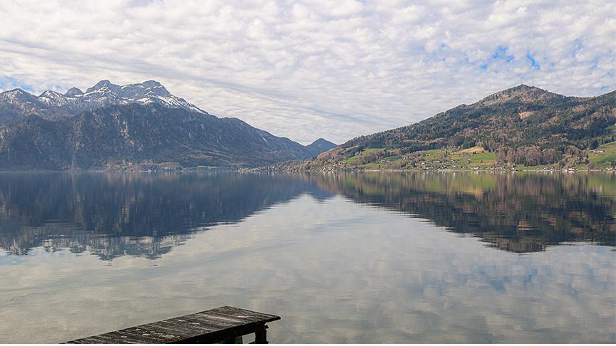 See mit Steg und umgeben von Hügeln und Bergen, am Himmel eine lose Wolkendecke auf vielen kleinen Wolken, die sich auch im See spiegeln