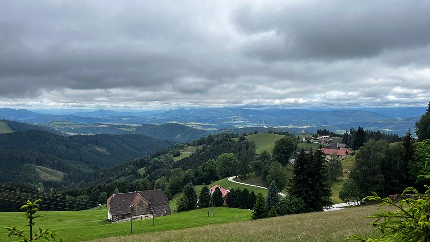 Hügelige Landschaft mit Häusern und wälden unter einer bauschigen Wolkendecke.