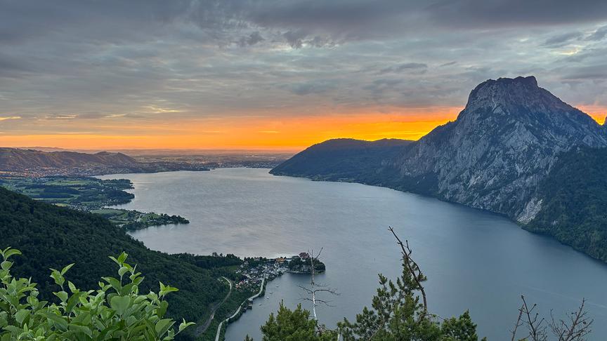 Von oben herab fotografierter See mit Bergen bei warm-orangem Sonnenaufgang unterhalb einer grauen, durchgängigen Wolkendecke.