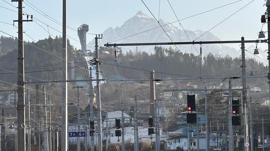 Durch die Oberleitungen des Bahnhofes aus, ist eine Skisprungschanze und ein Berg im Hintergrund zu sehen. Die Sicht ist leicht milchig.