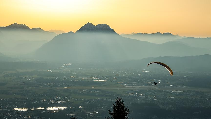 Tal mit Bergkulisse im Hintergrund ist etwas verschleiert durch Staub und wird daher von der Sonne in ein weiches Licht gehüllt, im Vordergrund ein Paraglider
