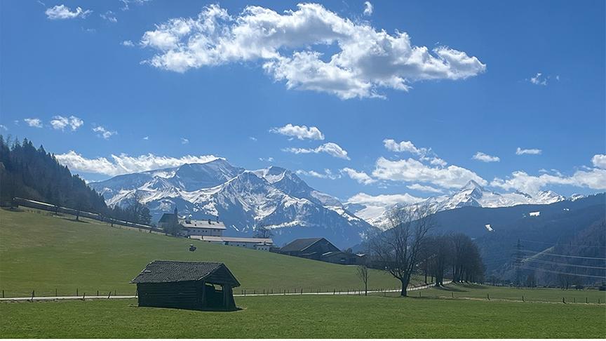 Grüne Weiden mit Häusern und Bäumen, im Hintergrund hohe, schneebedeckte Berge mit blauem Himmel und wenigen Wolken