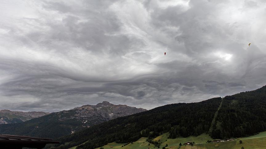 © Horst Fankhauser Panoramaaufnahme von einer bergigen Landschaft. De Berg im Vordergrund ist bewaldet, eine Schneise mit einem Lift zieht sich hinauf. Im Hintergrund sind mehrere felsige Berge zu sehen. Der Himmel ist düster und von einer geschwungenen Wolkendecke bedeckt. Zwei Paraglider fliegen am Himmel.