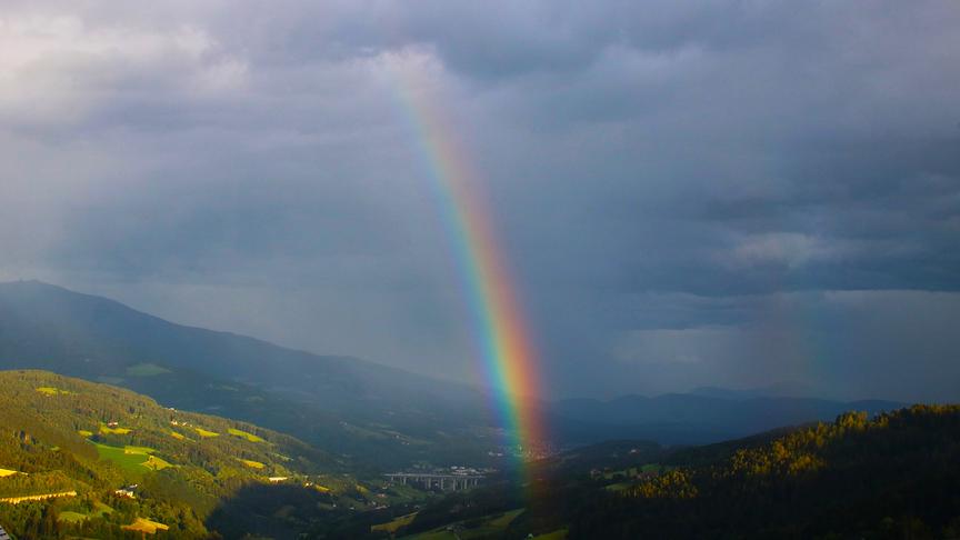 Regenbogen zwischen grauem, wolkenbehangen Himmel und der darunterliegenden leicht hügeligen, begrünten Landschaft mit Gebäuden. Links wird ein Teil der Landschaft bereits wieder von der Sonne beschienen.