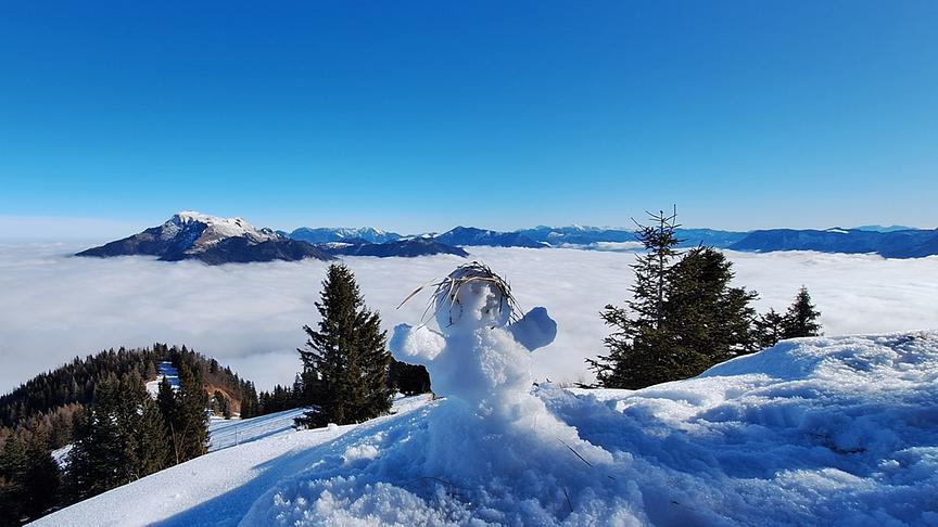 Im Vordergrund ist ein kleiner Schneemann, im Hintergrund sieht man von oben herab auf den Nebel, der zwischen den Bergen hängt.