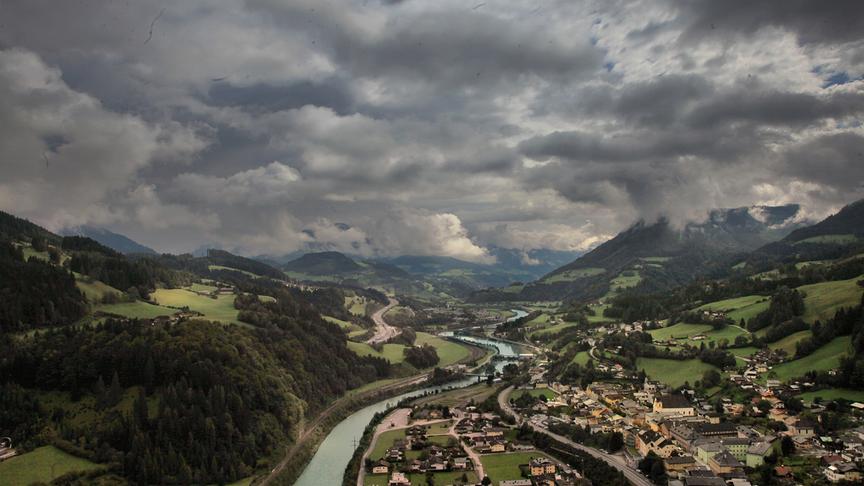 Luftaufnahme über ein Tal mit Siedlung, Fluss und Wäldern in einer hügeligen Landschaft unter hohen Wolken.