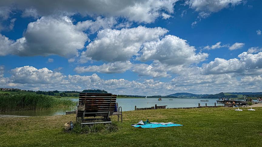 © Doris Stemeseder Blick vom grasigen Ufer über einen See und niedrige Hügel. Am Ufer liegt ein Handtuch und Bänke, ein Steg führt in der Ferne ins Wasser. Über der Szenerie sind zahlreiche bauschige Wolken, die für ein Muster an blauen Schönwetterhimmel sorgen.