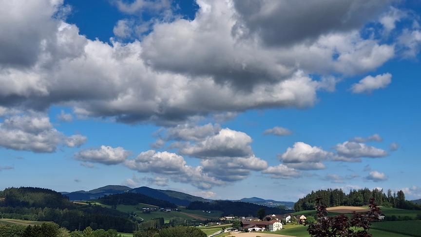 © Erwin Falkner Hügelige Landschaft mit Wiesen, Feldern, Wäldern und einigen Häusern, über die mehrere bauschige Wolken hinwegziehen am blauen Himmel.