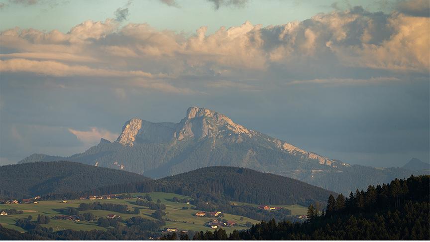 © Christoph Manuel Stockner Hügelige Landschaft mit Wiesen, Wäldern und vereinzelten Häusern. Im Hintergrund hohe, felise Berge und eine beeindruckende Wolkenformation, die milchig wie durch einen Schleier betrachtet wirken.