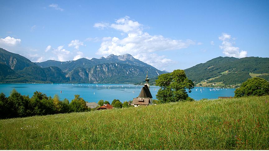 Ein türkisblauer See liegt zwischen Bergen und Wiesen. Einige Wassersportler und Boote sind am See. Am Ufer ist ein Dorf. Der Himmel ist blau mit einigen Wolken.