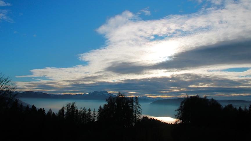 Aufziehende Wolken über Bergen, Bäumen und einem Gewässer.