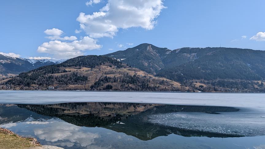 Der See ist nur noch wenig zugefroren, im Wasser spiegeln sich die Berge aus dem Hintergrund, blauer Himmel und eine Wolke
