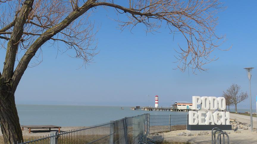 Bebautes Ufer eines Sees, im Vordergrund ein Schriftzug mit Podo Beach, im Hintergrund ein Steg mit Leuchtturm