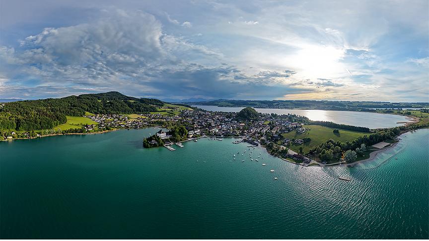 Bewölkter Sonnenschein über einem türkisfarbenen See und einer am Ufer liegenden Ortschaft.