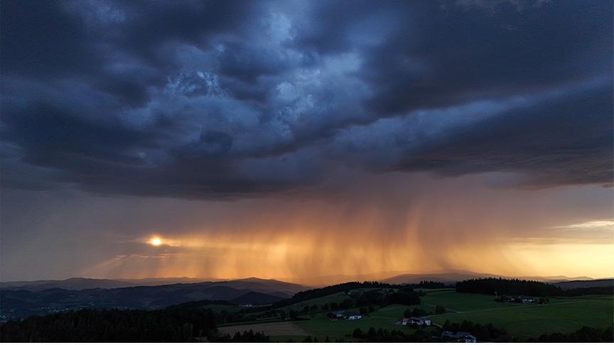 Eine dramatische Landschaftsaufnahme zeigt eine weite, hügelige Landschaft unter einem imposanten Gewitterhimmel. Über den sanften, grünen Hügeln und verstreuten Häusern im Vordergrund hängt eine dichte, dunkelblaue bis fast schwarze Wolkendecke. Am Horizont bricht im Hintergrund die tiefstehende Sonne durch eine Lücke, wodurch der dort fallende Starkregen in ein intensives, goldenes und orangefarbenes Licht getaucht wird. Die Regenschauer sind als deutliche, vertikale Streifen sichtbar, die wie ein leuchtender Vorhang den dunklen Himmel mit der erleuchteten Erde verbinden.