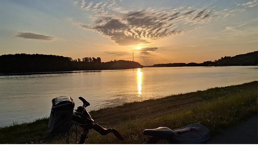 Sonnenaufgang über der Donau. Im Vordergrund ist ein Fahrrad neben dem Fluss abgestellt. Über den Himmel ziehen sich wellenartige Wolken.