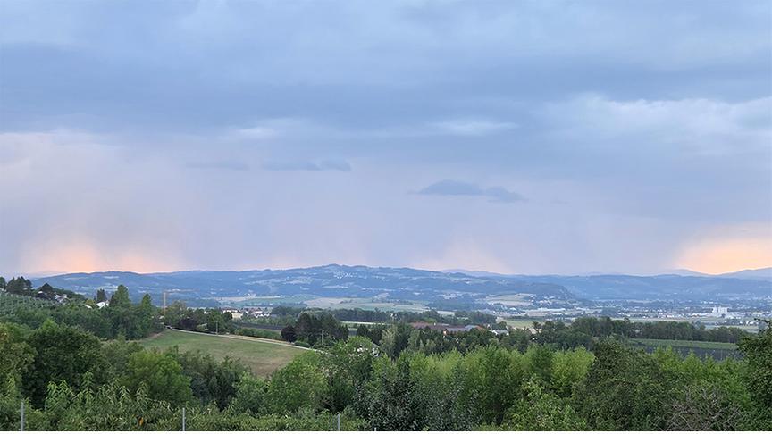 Weite Aufnahme einer leicht hügeligen Landschaft, über der sich graue Wolken erstrecken und Regenschleier sich gen Boden ziehen.