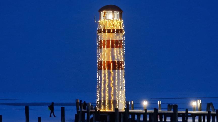 In der Dunkelheit von Lichterketter hell-erleuchteter Leuchtturm. Im Hintergrund die Schemen einer Person, die auf dem zugefrorenen See spazieren geht.