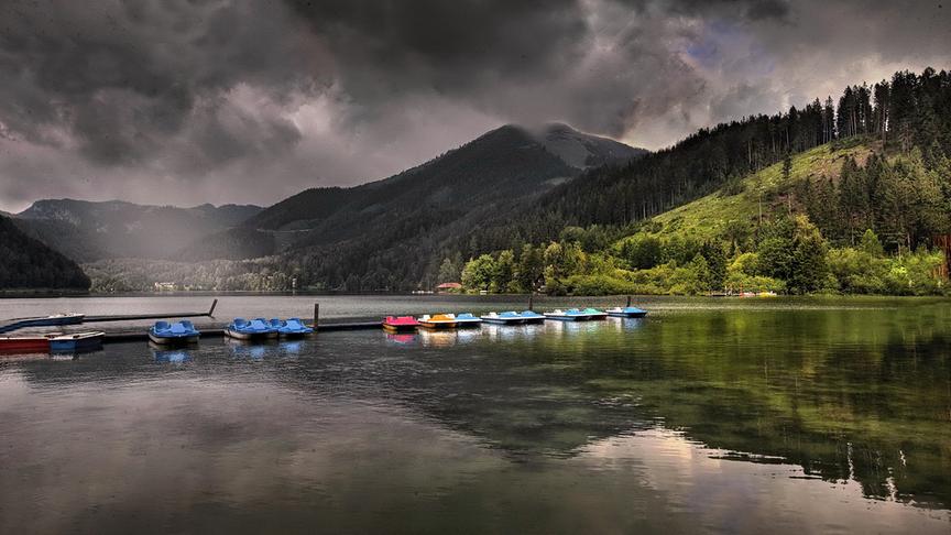 Dunklte Stimmung und Wolken über einem See umgeben von Bergen. Die Tretboote am Steg bilden einen farbigen Kontrast.