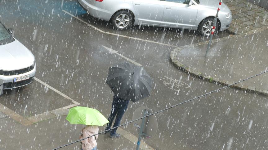Zwei Personen mit Regenschirmen auf einer Straße, dicke Regentropfen fallen vom Himmel.