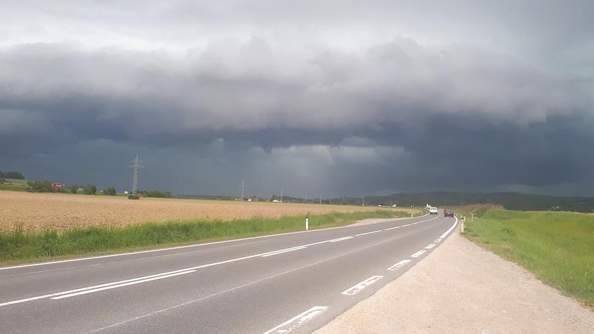 Eine Straße zwischen einem Feld und einer Wiese bei dunklen Wolken.